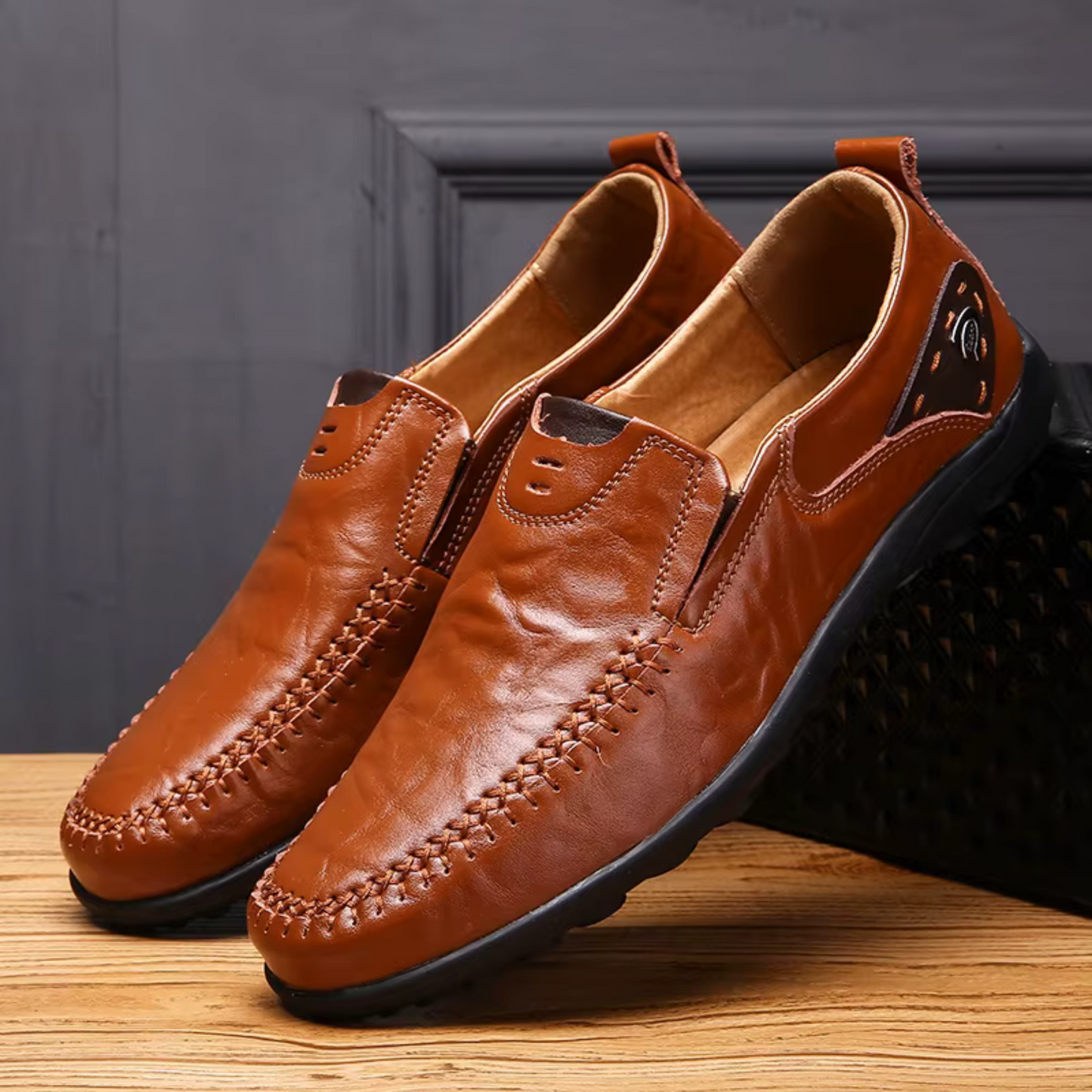 Brown leather slip-on moccasins with detailed stitching and black rubber soles on wooden surface